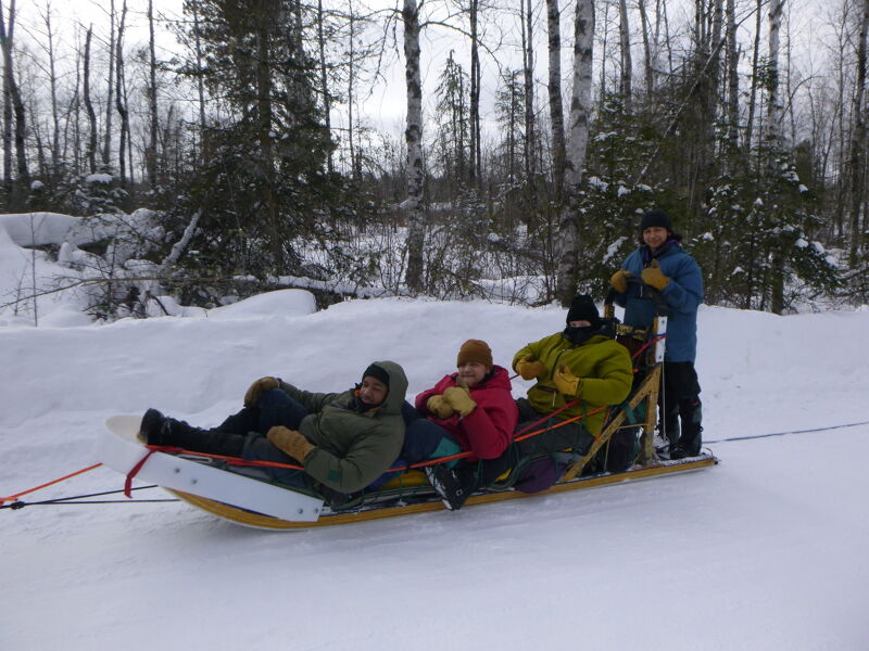 The image shows a group of people on a sled in a snowy environment. There are four people on the sled, and one person is standing behind it. They are all wearing winter clothing. The background consists of trees and snow.
